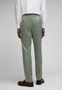 Hellegrüne, maßgeschneiderte Hose aus strukturiertem Stoff, mit zwei rückseitigen Taschen mit Knöpfen, kombiniert mit braunen Schuhen.