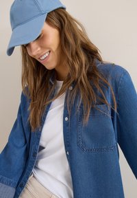 Femme portant une casquette bleu clair, une chemise en jean ample par-dessus un t-shirt blanc, souriante et regardant vers le bas sur un fond uni.