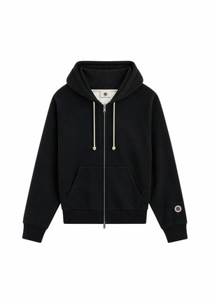 Sudadera negra con cremallera, bolsillos delanteros, cordones blancos, puños de canalé y un pequeño parche circular en la manga izquierda cerca de la muñeca.