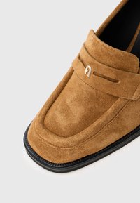 Mocassino in suede marrone con suola nera e un piccolo dettaglio logo metallico sul cinturino anteriore, mostrato su uno sfondo bianco.