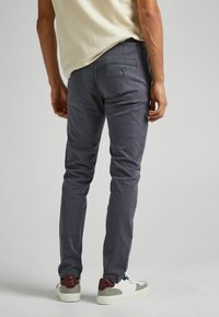 Mężczyzna w szarych dopasowanych spodniach, białych trampkach z bordowymi i szarymi akcentami oraz w jasnobeżowej koszuli z krótkim rękawem, stojący na jednolitym tle.