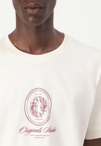 Cremefarvet bomulds T-shirt med et cirkulært rødt grafik, der har indviklede designs og tekst, samt en ribbet halskant og korte ærmer.