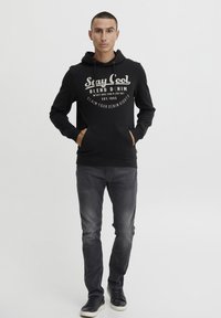 Sudadera negra con impresión blanca que presenta el texto "Stay Cool", combinada con jeans gris oscuro y zapatillas negras. Tejido suave, capucha con cordón.