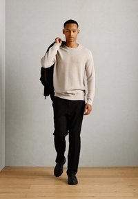 Pull en tricot beige clair, pantalons noirs, chaussures noires. Le modèle tient une veste noire, marchant sur un sol en bois contre un fond gris.