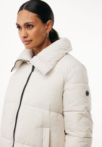 Giacca imbottita color beige chiaro con colletto alto, zip nera, due tasche frontali e accento del logo sulla manica sinistra.