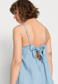 Top de alças ajustáveis em chambray azul claro, com detalhe de laço nas costas e um recorte em forma de olho-chave. Textura suave e design fluido.