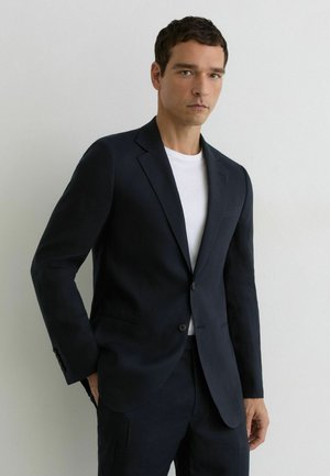 Hombre vestido con una chaqueta y pantalones de traje negros y una camiseta blanca, de pie contra una pared blanca lisa, con las manos parcialmente en los bolsillos.