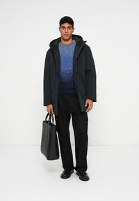 Μαύρο επενδυμένο παλτό με κουκούλα, μπλε gradient πουλόβερ, μαύρο παντελόνι και μαύρα παπούτσια. Το μοντέλο κρατά μια μαύρη τσάντα tote. Απλός φόντος.