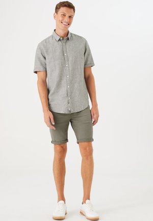 Chemise à manches courtes grise avec boutons, shorts avec revers verts, et baskets blanches. Le tissu semble léger et a une coupe décontractée.