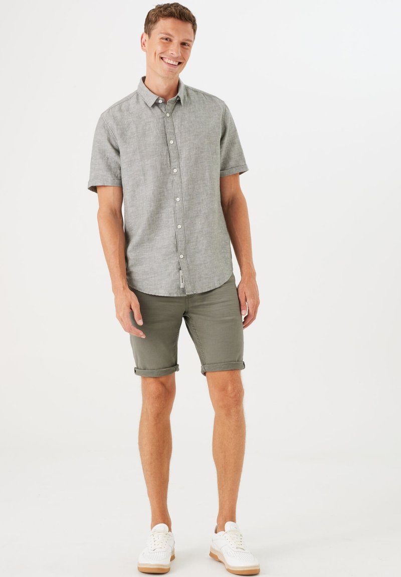 Camisa de botones de manga corta gris, pantalones cortos verdes con dobladillos enrollados y zapatillas blancas. La tela parece ligera y tiene un ajuste casual.