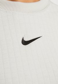 Ljusgrå Nike sweatshirt med ribbad krage, texturerad tyg och en svart broderad swoosh-logotyp. Vertikal ribbad mönster synlig.