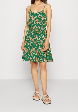 Mujer con vestido verde de flores hasta la rodilla, con tirantes finos y sandalias beige con doble hebilla, de pie contra un fondo blanco liso.