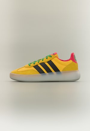 Žuta tenisica s crnim prugama, zelenim vezicama, ružičastim unutarnjim oblogom i prozirnim potplatom, prikazana sa strane na jednobojnoj pozadini.