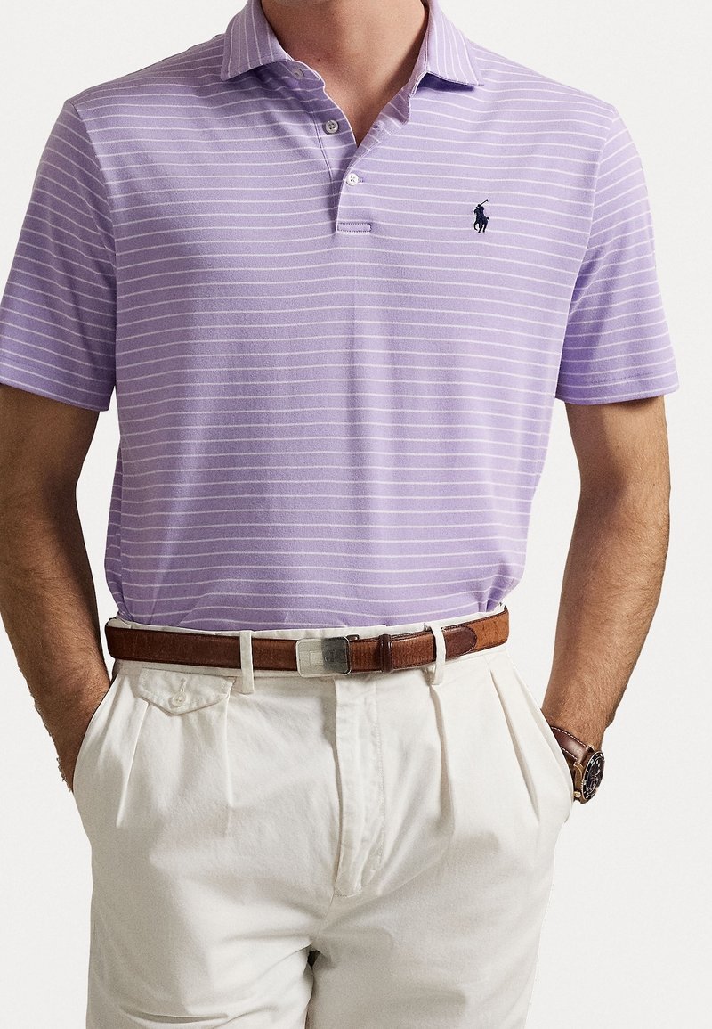 Uomo che indossa una polo a righe lavanda infilata in pantaloni bianchi plissettati con una cintura in pelle marrone e un orologio da polso.