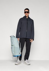Dunkelgraue wasserdichte Jacke mit Kapuze, über schwarze Hosen getragen. Hellblauer Rucksack mit grünen Akzenten in einer Hand gehalten. Weiße Sneaker.