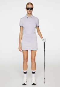 Ljusgrå polo tröja med korta ärmar, matchat med en figurnära grå kjol. Modellen bär svarta solglasögon och håller en golfklubba, iklädd vita strumpor.