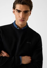 Schwarzer Sweatshirt mit Struktur und Rundhalsausschnitt, mit dem "GUESS"-Logo. Getragen über einem blauen Hemd mit Kragen und Knöpfen am Hals.