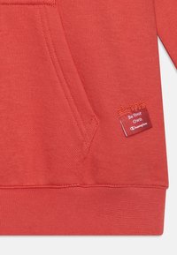 Röd hoodie med texturerad tyg, känguruficka, ribbad nederkant och en märkeslapp som detaljerar "Be Your Own" med en Champion-logotyp.