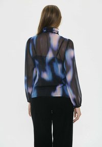 Caurspīdīga blūze ar garām piedurknēm zilā un violetā ombrē rakstā, ar kraukšķīgu apkakli un pieguļošām aprocēm, apvienota ar melniem biksēm.