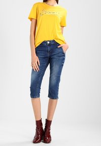 Mujer vestida con una camiseta amarilla con un gráfico, unos pantalones vaqueros de denim azules hasta la rodilla y brillantes botas rojas hasta el tobillo, de pie con una mano en el bolsillo.