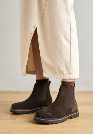 Bottines en daim marron portées avec des chaussettes marron et une jupe beige avec une fente devant, debout sur un sol en bois.