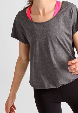 Kvinde iført en løs grå t-shirt med korte ærmer over en knaldrød sports-bh og sorte leggings mod en ensfarvet lys baggrund.