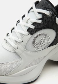 Baskets Michael Kors blanches et noires avec lacets blancs, panneaux texturés et logo MK sur le côté près du col de la cheville.