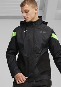 Zwarte windbreakerjas met groene accenten, netvormige zijpanelen en een ritssluiting aan de voorkant. Logo-details zijn op de borst en schouder aanwezig.