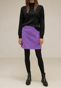 Musta tekstuuriga kudumislest ja lilla A-linja mini seelik, mis on varustatud kahe esirauvaga, koos mustade pahkluu saapade ja tumedate sukkpükstega.
