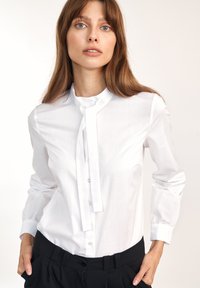 Biała bluzka z długim rękawem z kołnierzykiem mandarynkowym, z przodu z guzikami oraz wiązaniem przy dekolcie. Materiał wydaje się gładki i lekki.