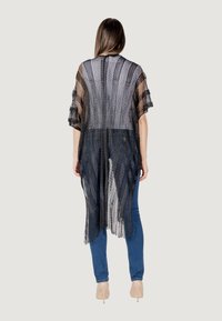 Kimono lungo nero con frange, realizzato in pizzo a rete, caratterizzato da un design aperto e maniche larghe, indossato sopra jeans blu.