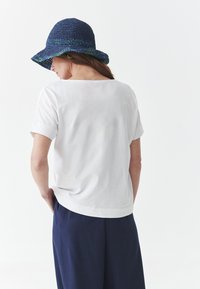 Chemise blanche à manches courtes en tissu doux, associée à un pantalon bleu marine ample et un chapeau de soleil bleu tressé à large bord.