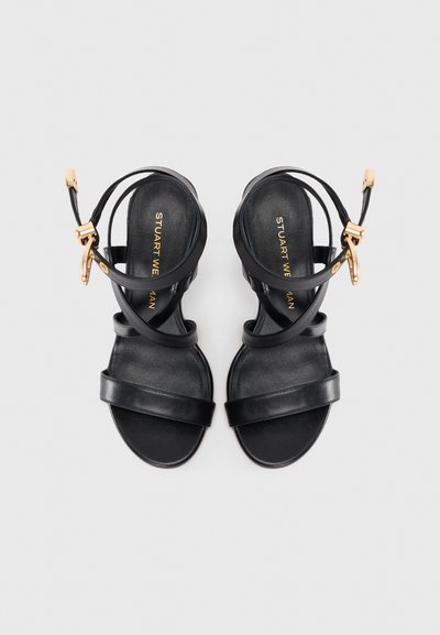 Een paar zwarte leren platte sandalen met enkelbanden en gouden gespen op een witte achtergrond.