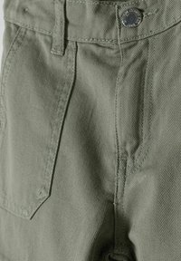 Pantalones cortos de algodón verdes con textura desgastada, que cuentan con un bolsillo frontal, lazos para cinturón y un cierre de botón metálico. Costuras visibles a lo largo de las costuras.