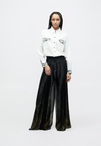 Valkoinen napillinen paita, jossa seepra-kuvioidut taskut, yhdistetty mustiin, laskostettuihin leveälahkeisiin housuihin, joissa on hienovarainen kultainen kuvio.