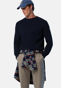 Jersey de punto acanalado azul marino combinado con una camisa de franela a cuadros atada a la cintura, usado con pantalones cargo caqui y una gorra de color azul claro.