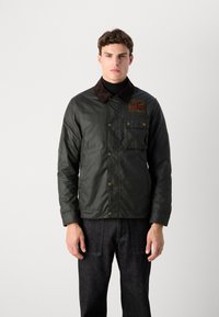 Veste en coton ciré vert foncé avec un col en velours côtelé marron, une poche avant et un logo brodé sur la poitrine.