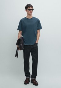 Maglietta di cotone verde petrolio scuro con collo rotondo, abbinata a jeans scuri e scarpe stringate marroni. Una borsa nera in pelle è tenuta in una mano.