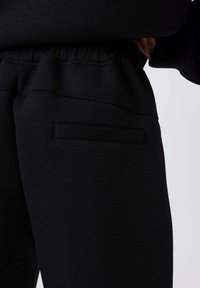 Zwarte joggingbroek met een gladde textuur, elastische tailleband en een plat zakdetail aan de linkerkant. Het ontwerp heeft eenvoudige lijnen.