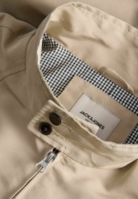 Γιακάς μπεζ σακακιού με επένδυση σε ασπρόμαυρο καρό, δύο σκοτεινά κουμπιά και ασημένιο φερμουάρ, που δείχνει την ετικέτα της μάρκας Jack & Jones στο εσωτερικό.