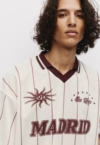 Weißes Mesh-Jersey mit kastanienbraunen Streifen und Akzenten. Mit einem Sonnenmotiv auf der linken Seite und "MADRID" in Fettdruck. Klassisches Kragen-Design.