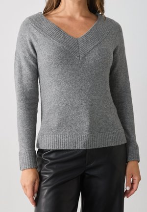 Femme portant un pull en maille gris à col en V et un pantalon en cuir noir, visible du cou aux hanches sur un fond uni.