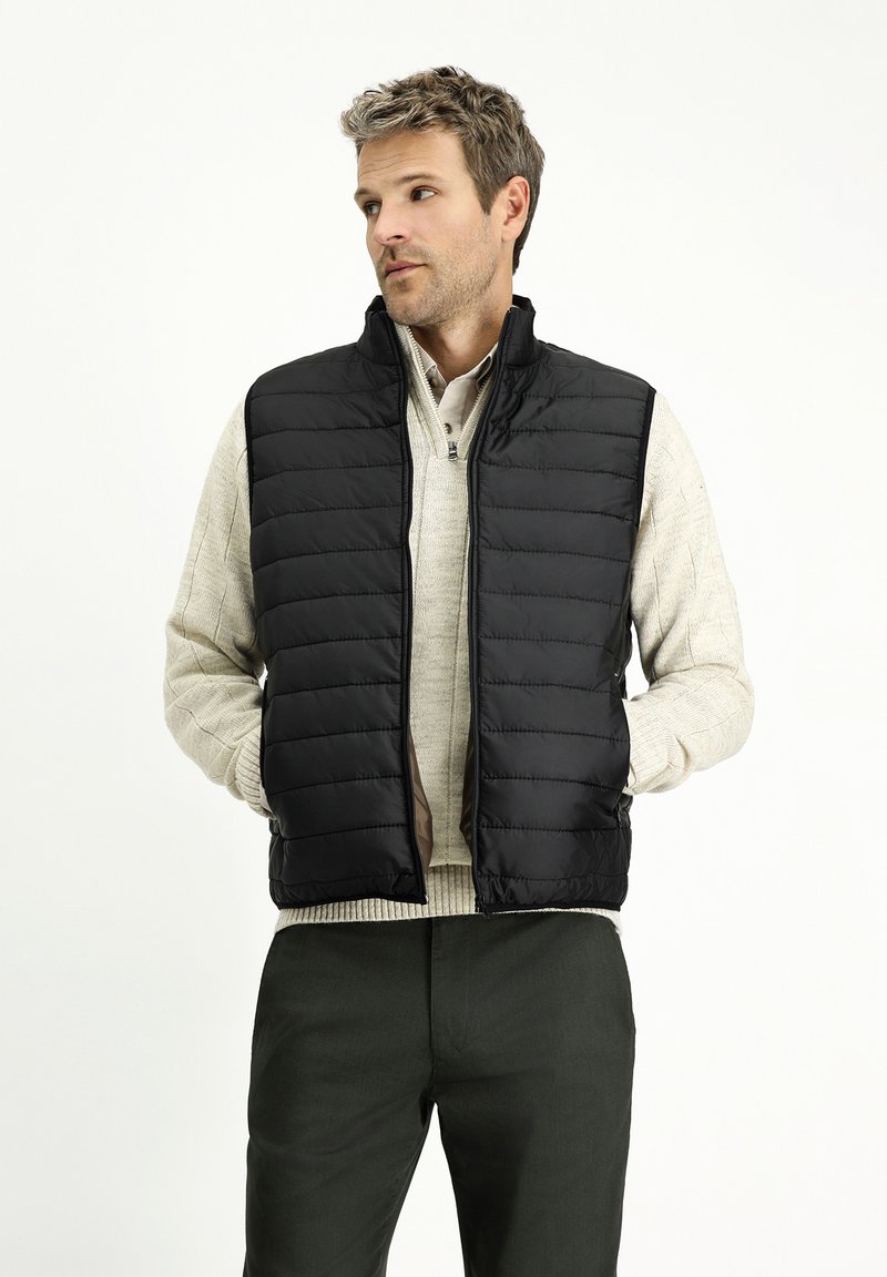 Gilet imbottito nero con colletto alto, dotato di chiusura con zip e texture a trapunta orizzontale, indossato sopra un pullover beige chiaro.