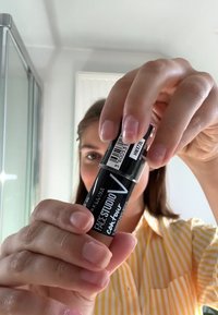 Svart och klart förpackning med en vridbar design, märkt "FACE STUDIO V CONTour," hålls i en hand ovanför en randig gul skjorta.