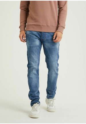 Mand iført falmede blå jeans, hvide sneakers og en brun langærmet top, stående mod en enkel lys baggrund.