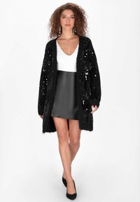 Czarny kardigan z cekinami o puszystej fakturze, z otwartym przodem, połączony z dopasowaną czarną skórzaną spódnicą i białą bluzką z głębokim dekoltem w kształcie litery V.