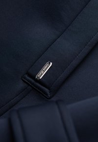 Primer plano de una tela azul marino que presenta una etiqueta cosida con "Oliver Spencer" en acabado metálico y un pequeño agujero para fijación. Textura suave.