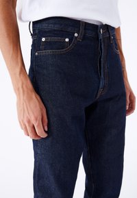 Jeans de mezclilla azul oscuro con un diseño de pierna recta, con costuras naranjas, estilo de cinco bolsillos y cierre de cremallera con botón.