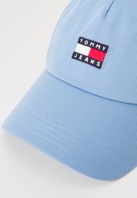 Blå baseballcap lavet af struktureret stof. Har et broderet logotag med røde, hvide og blå accenter. Buet skygge og justerbar pasform.