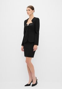 Blazer noir ajusté avec un décolleté profond, trois boutons et des épaules accentuées, associé à une jupe noire ajustée et des escarpins noirs élégants.